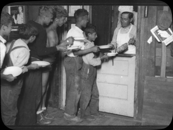 Group of boys standing in line, holding empty plates to be filled by a cook, wearing a chef's hat, who is standing behind a half-door with a pan in one hand and a plate in the other. The settlement house sent some area children to camp in the summer.