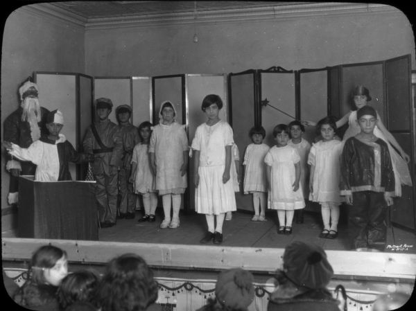 Performers lined up on the stage in "The Toy Shop," a Christmas play, with a Santa figure, a boy in harlequin dress, two boys in military uniforms, girls in filmy white dresses, a girl with a crown and a star wand, and a boy in Chinese-style clothing. Heads of audience members in the front row are in the foreground.