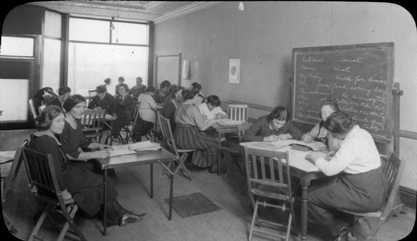Group of women seated at tables with a freestanding blackboard on which are written English words and phrases. One of the women in the foreground appears to be looking at the camera.