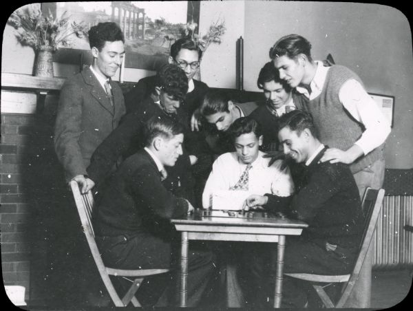 Young men playing a game of checkers at Neighborhood House. Three men are seated at a small table with the game board, while six others crowd around them, looking on.