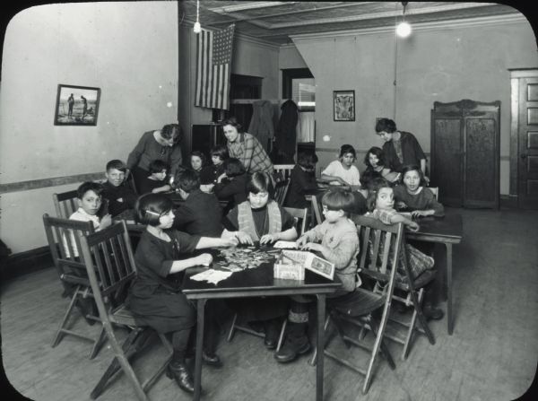 Children seated at tables playing board games and working on a jigsaw puzzle, with three women assisting.