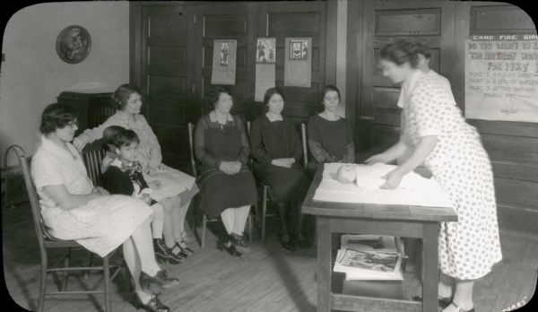 View of a woman standing at a table demonstrating infant care (swaddling?) using a baby doll to five women and two children who are sitting in chairs facing her. Another woman is standing nearby, looking on. In the background, a hand-lettered notice addressed to Camp Fire Girls advertises how to earn points toward a birthday honor.