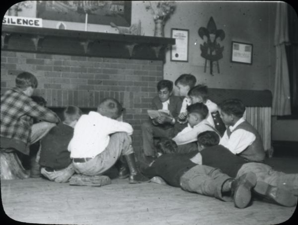 One boy reads a book to a group of boys who are seated or lying in front of him by a fireplace. A sign on the mantel reads, "Silence". Boy Scout insignia and other framed items are hanging on the wall.
