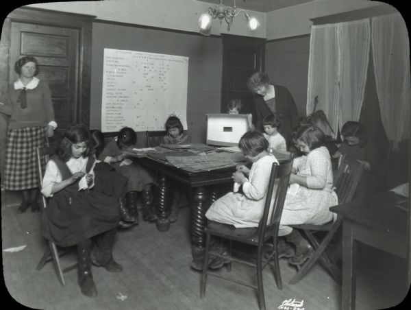 Image from an album kept by Neighborhood House, with girls sitting at a table sewing, Mary Lee Griggs standing and helping, and another woman standing by the door.
