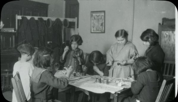 Group of girls sitting or standing around a table that is covered with paper and containers of paint, holding brushes and small objects they are painting. One girl is in a Camp Fire Girl uniform. A coat rack in the background is hung with caps and jackets.