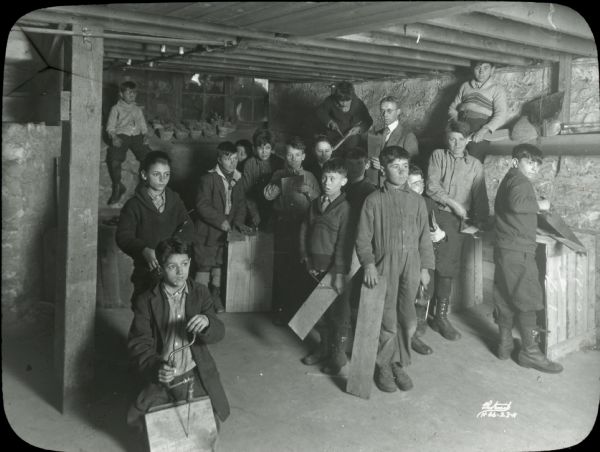 Group of boys at a woodworking session in the basement shop at Neighborhood House, with boys posing with various tools and planks of wood. In the far background a boy is sitting on the window ledge, and a crowd of boys is outside peering through the windows. On the right is a man standing in a suit coat holding papers (plans?).