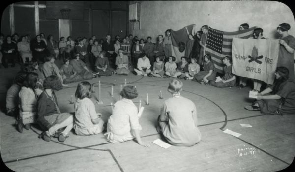 Group of Camp Fire Girls, some in uniform, sitting on the floor in a circle around lighted candles. Some girls are standing and holding Camp Fire Girls banners and an American flag. Adults and other children, including some Boy Scouts, are sitting on chairs in the background.