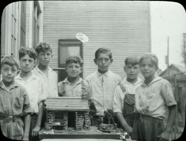Seven boys standing outdoors around a table with a model of a log cabin and its surroundings on it. A building with narrow siding is in the background.