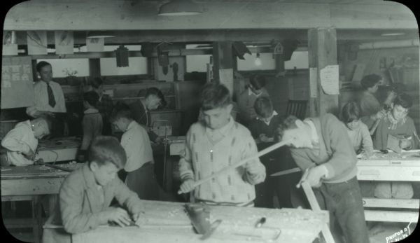 Group of boys working on various woodworking projects: sawing, hammering, and marking. A man in a shirt and tie is talking to one of the boys in the background. Birdhouses hang from the rafters, and other projects, such as wooden animals, are displayed on shelves.
