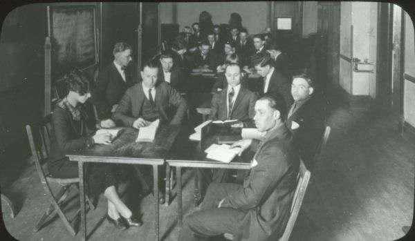 Groups of men, most dressed in suits and ties, sitting at small tables, looking at books. A few female teachers, including one in the foreground, are also present. A blackboard stands off to the side.