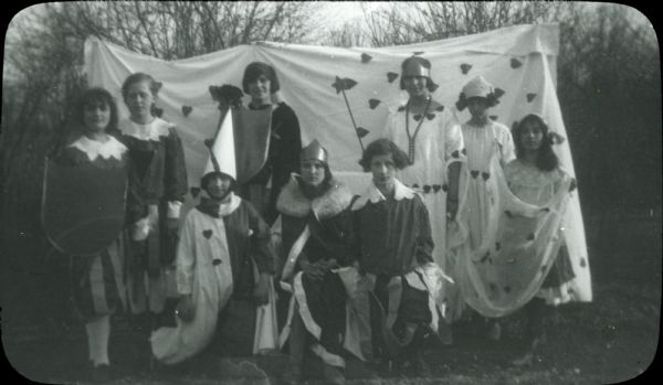 Cast members of a play staged for Valentine's Day, with costumed figures holding shields, a jester in a half-white and half-black costume, a woman with a crown and a fur-collared cloak, and a woman wearing a crown and two girls in white dresses decorated with hearts. A backdrop decorated with hearts is behind them.