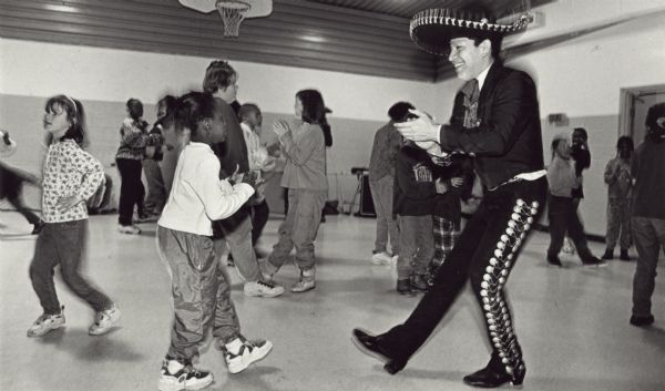 Jesus Avila of Ballet Folklorico in a sombrero and suit with side-seam decoration on the trousers claps and dances with children in a gymnasium, probably at Neighborhood House.