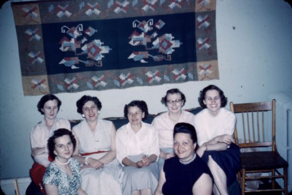Group of Ladies' Sewing Club women posing under a tapestry at Neighborhood House.