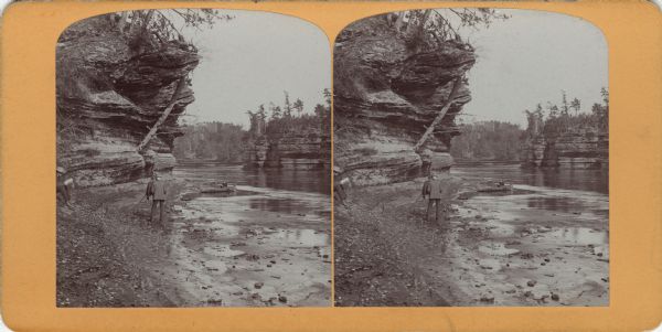 Stereograph of H.H. Bennett manning his camera on shore near Steamboat Rock. A woman is in a canoe near the shoreline, and a man is reading a book on the far left.