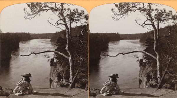 Ruth or Miriam Bennett in a plaid dress and hat sitting on top of High Rock. Chimney Rock is located below, on the right side of the river.