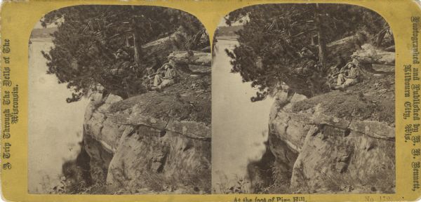 Stereograph of three unidentified men laying on the rock formation over the river. Text at left: "A Trip Through the Dells of the Wisconsin."