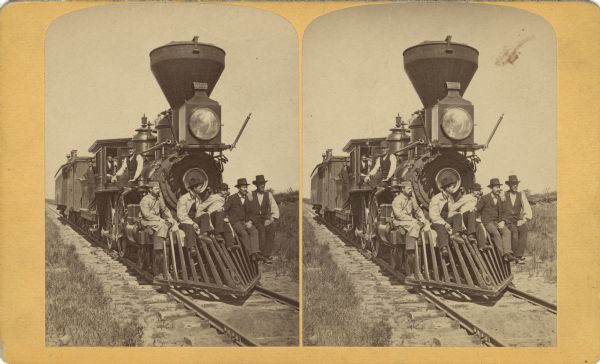 Men posed on the front cowcatcher, or pilot, of a wood burning locomotive, with a few cars attached. Three other men pose on the locomotive.
