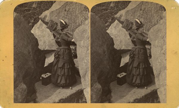 Stereograph of Mrs. A.S. Richards posing with a camera and tripod over her shoulder among boulders. There is a box, probably holding glass-plate negatives, at her feet.