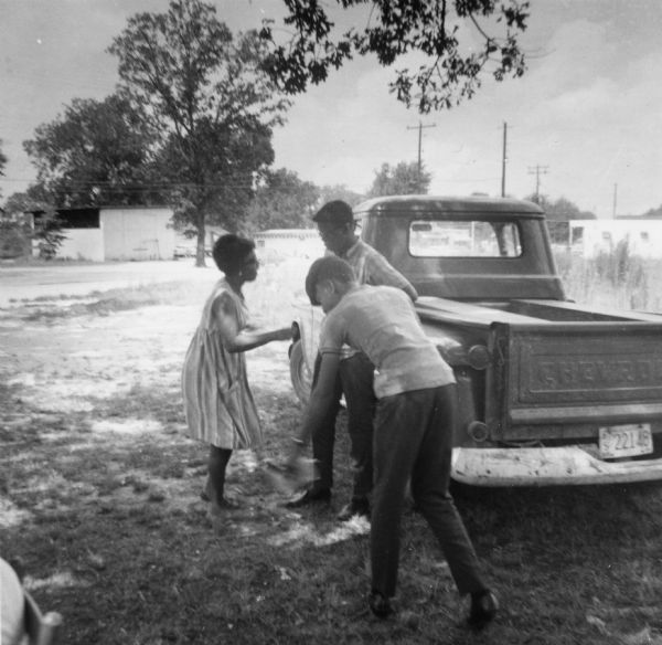 Three people stand outdoors near a truck acting out a group improvisation of a slave revolt. They were attending Freedom School during Freedom Summer at Palmer's Crossing, near Hattiesburg. "Acting out history was a popular activity in the Freedom School curriculum. The student in center is the 'master'; the two others are his faithful 'toms' betraying a plot by fellow slaves to him. The students liked to play the role of 'tom' to get out their anger at those who played that role in real life."