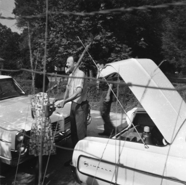 View through fence of Pete Seeger and an unidentified woman stand outdoors near two parked cars. He was there for a concert he played in a church during Freedom Summer. Another man stands in the road behind him.