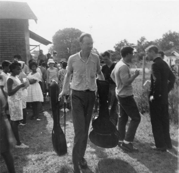 Pete Seeger walking outdoors through a crowd carrying two instrument cases. The concert took place during Freedom Summer near Hattiesburg. "Pete Seeger sang inside a church where the temperature rose above 100 degrees."