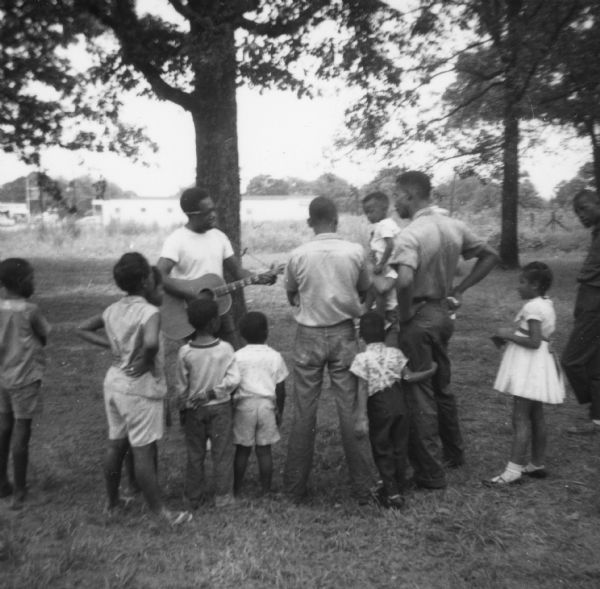 A group of adults and children at an impromptu concert held outdoors during Freedom Summer outside of the Freedom School at Priest Creek Baptist Church.  Likely participating in music workshops that were a part of the Mississippi Summer Caravan of Music.