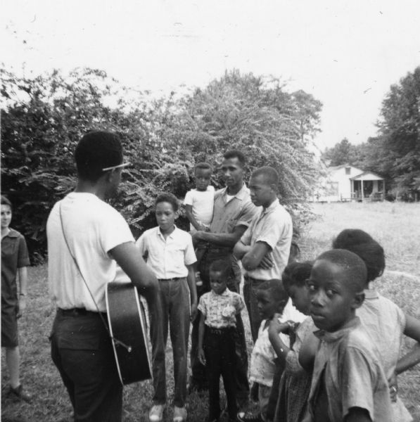 A group of adults and children at an impromptu concert held outdoors during Freedom Summer outside of the Freedom School at Priest Creek Baptist Church. Likely participating in music workshops that were a part of the Mississippi Summer Caravan of Music.