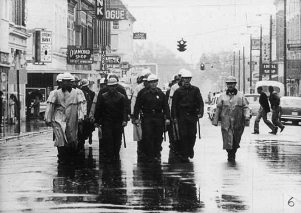 A group of Hattiesburg police officers wearing helmets and carrying batons walk in formation down a street in the rain at a Freedom Day on their way to Forrest County Courthouse where African Americans protest that they have had their right to vote denied. Photo credits Danny Lyon.