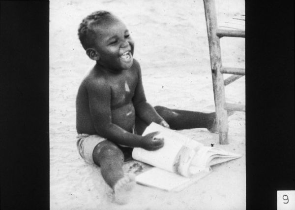 A toddler sits on the ground and laughs while paging through a book.