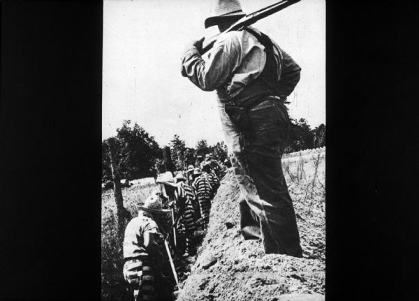 A group of correctional inmates work in a trench, while a man with a firearm watches from above.