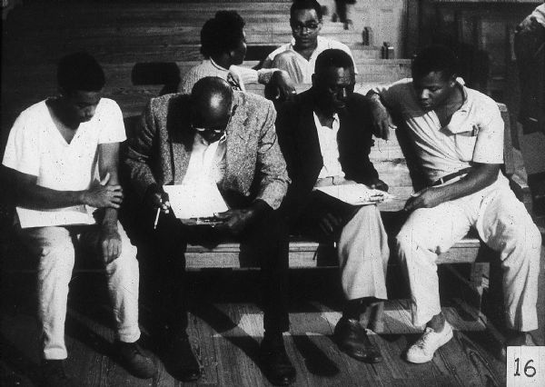 Four men sit on pews examining paper on clipboards. Behind them, Bob Moses talks with a woman.