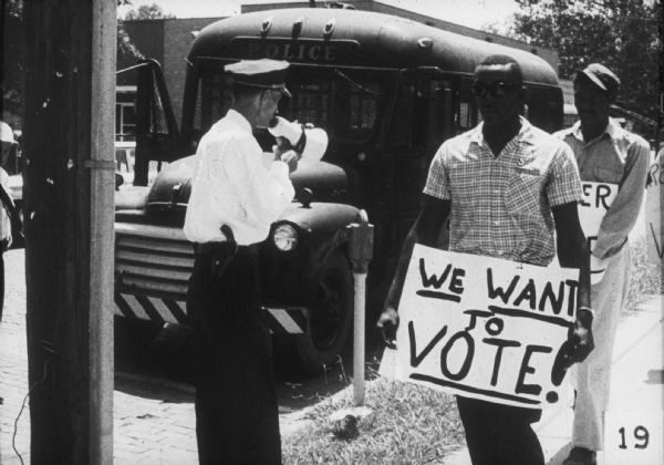 Two men holding protest signs are walking down a sidewalk past a white police officer who is holding a bullhorn at a Greenwood Freedom Day. In the background, a police vehicle is parked on the curb.