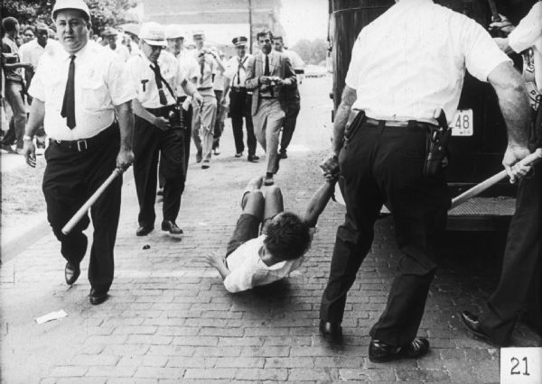 Police drag away 15-year-old Annie Lee Turner. Officers are wearing helmets and holding batons. A couple of men with cameras are walking towards the person on the ground.