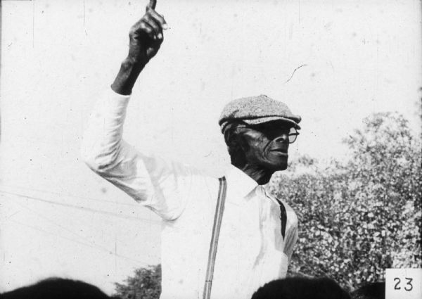 A man stands with his arm raised and his finger pointed upward at a mass meeting.