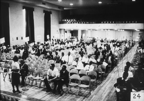 A group of people are seated on folding chairs, likely at the MFDP state convention. A woman at the front of the room addresses the group through a microphone.