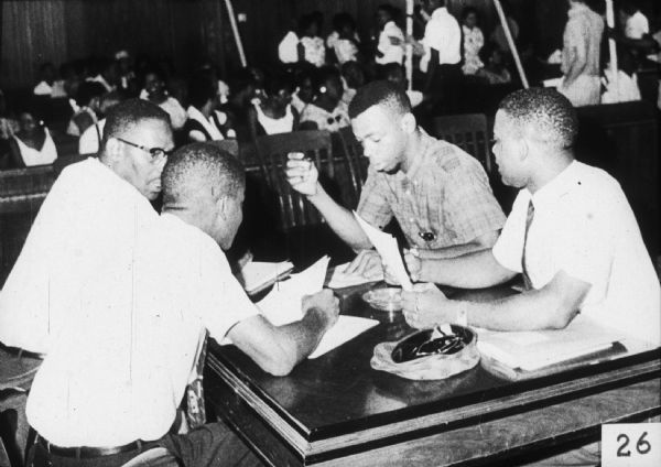A group of four African-American men are seated at a table looking at papers. Behind them a large group of people are sitting and standing.