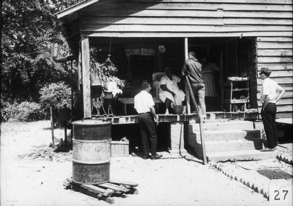 Two women stand on the porch of a home. Three men, probably Freedom Summer volunteers, are talking to them.