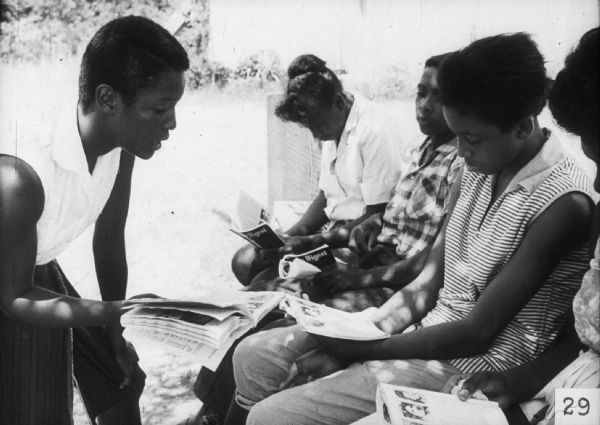 A group of women and girls seated outdoors read copies of the same magazine, possibly as part of a Freedom School lesson.