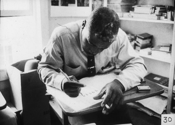 A man is seated at a desk, writing and smoking a cigarette.