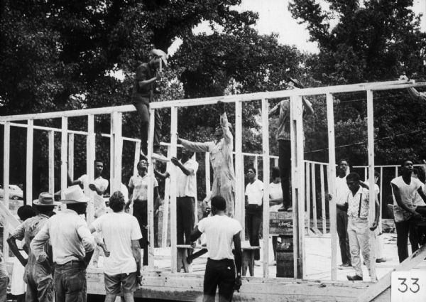 A group of men and women work on a community construction project. Several support the framing of a wall, while a man straddles the top of the frame to hammer in nails.
