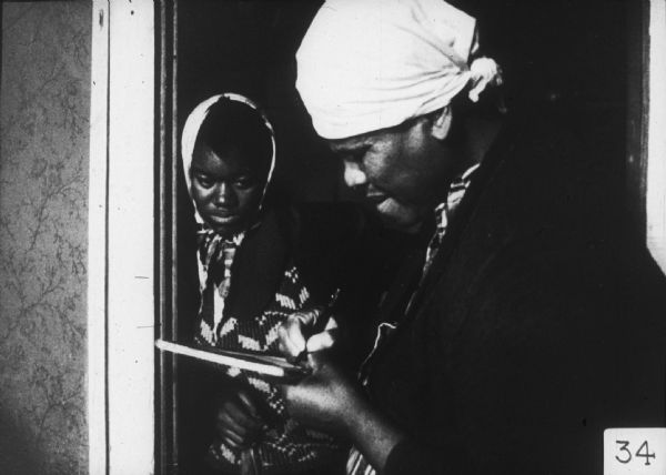 A woman in the foreground writes on a pad while a young woman looks on from a doorway.