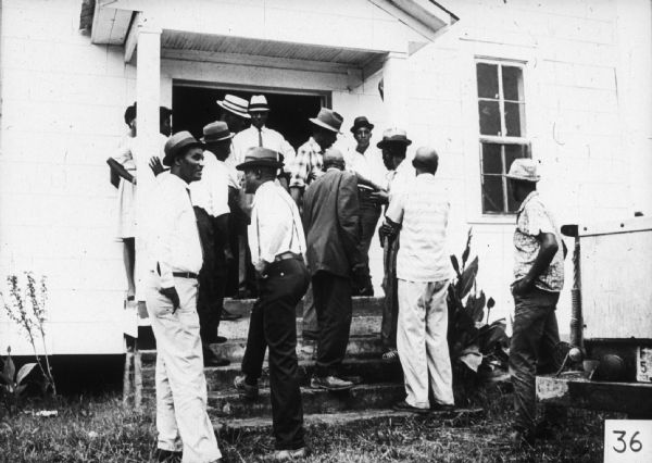 A group of men and women talk outside on the steps of a building and in the open doorway likely attending a mass meeting.