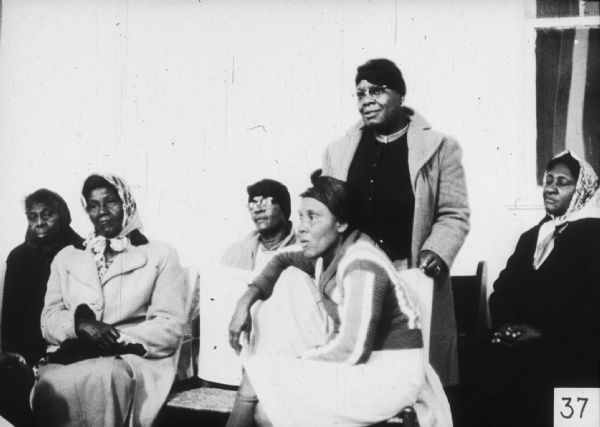 Six women, all wearing kerchiefs on their heads, are seated in a group. One woman is standing.