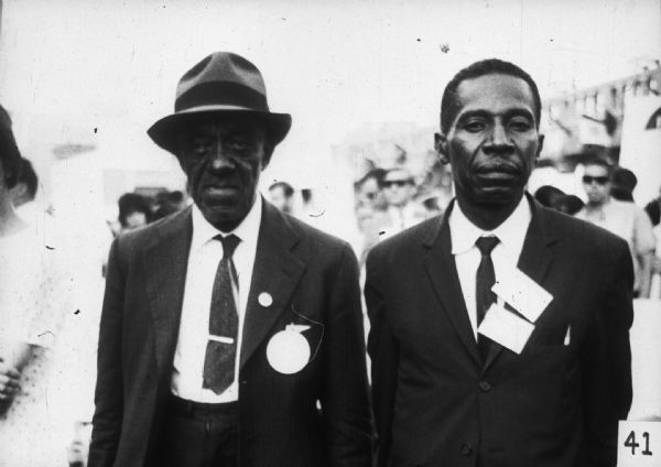 Two men wearing suits stand in front of a crowd of people.