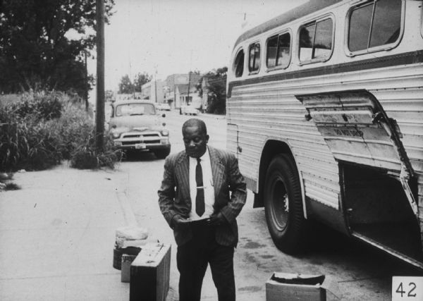 A man stands outdoors next to a motorcoach with an open door. There is luggage on the curb next to him.