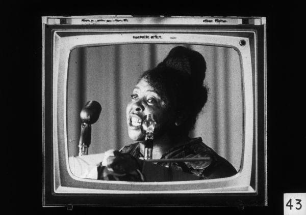A television set displays an image of Fannie Lou Hamer, civil rights activist and political figure, speaking into microphones.
