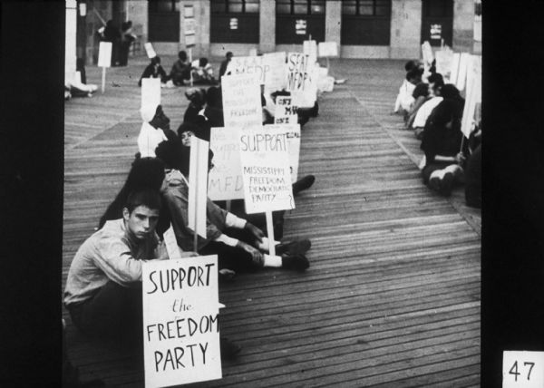 An integrated group of mostly young people sitting on the Atlantic City boardwalk. Many of them are holding signs showing support for the Mississippi Freedom Democratic Party (MFDP).