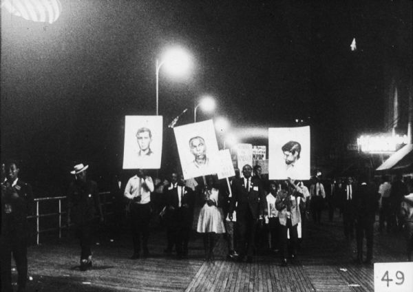 A nighttime rally on the Atlantic City boardwalk in support of the Mississipi Freedom Democratic Party (MFDP). Three people in front of the group carry signs with portraits of James Earl Chaney, Andrew Goodman, and Michael "Mickey" Schwerner, three civil rights volunteers who were murdered in the summer of 1964.