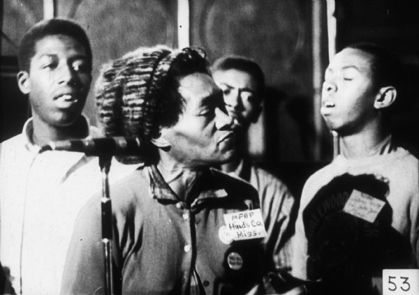 A woman wearing a knit cap stands in front of a microphone. The badge on her chest identifies her as an MFDP member from Hinds County. Three men behind her appear to be singing.