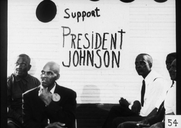 Several men sit in front of a large sign supporting Lyndon B. Johnson's 1964 presidential campaign.